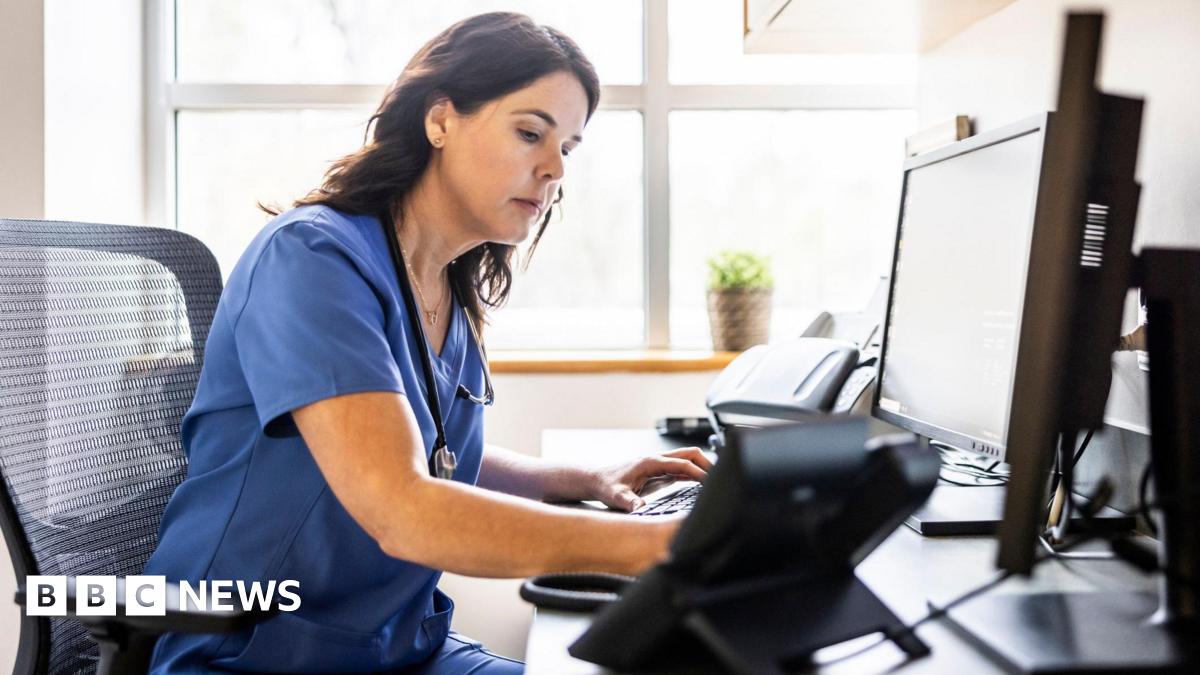 Nurse with long brown hair and blue scrub outfit works at a computer