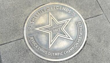 A gold-bronze coloured, circular metal plaque on the paving stone floor. Around the edge it reads "Sheffield Legends - Jessica Ennis Olympic champion heptathlete". In the centre is a star, with "London 2012" written in the middle of it.