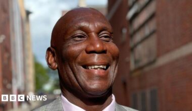 Uriah Rennie, a man wearing a grey jacket, lilac shirt and purple tie. He is smiling and appears to be walking down a narrow street between two large redbrick buildings.