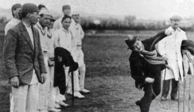 A black and white photo of a man wearing a coat, scarf and hat bowling a cricket bowl. A group of men are stood to the left if the photo in cricket whites.