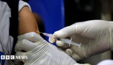 A person wearing white medical gloves injects a needle containing a clear, colourless vaccine into a child's arm.