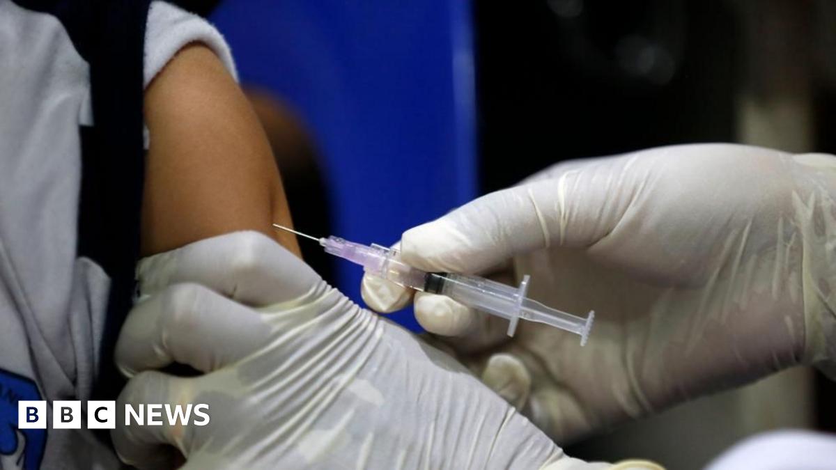 A person wearing white medical gloves injects a needle containing a clear, colourless vaccine into a child's arm.
