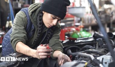 A woman with long black hair in two plaits wearing a black beanie hat, khaki hoodie and navy quilted bodywarmer working as a mechanic with her oil covered hands inside the engine of a car with its bonnet up