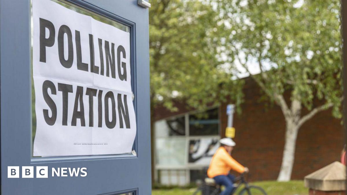 A polling station poster on the glass of an open door.- it reads "Polling Station" in black font on a white background. In the background there is a cyclist that is out of focus. They are wearing an orange coat and blue trousers and are cycling past two trees and a green lawn.