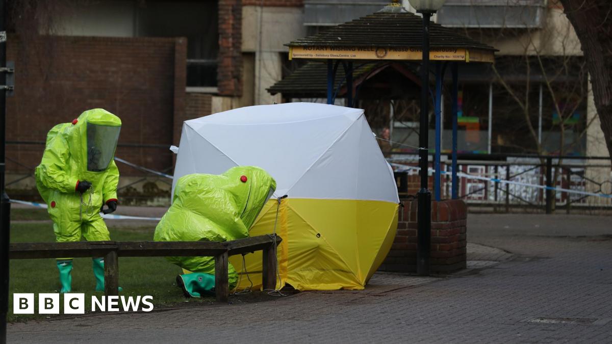 Two workers in bright yellow Hazmat suits crouch next to a forensic tent over a bench in Salisbury city centre