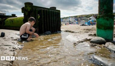 A boy in his shorts crouches in a stream of water on a beach next to a large mossy pipe with a grill on the end. In the distance a beach tent can be seen with people on the sand.
