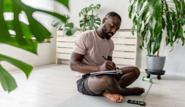 Young African-American man sitting cross-legged on a yoga mat at home, writing in a notebook after a workout, with a smartphone nearby, creating a calm and focused atmosphere