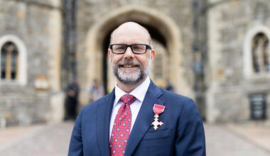 Portrait of Ken Henderson wearing a blue blazer and red tie standing outside a historic red building. On his lapel is a gold medal.