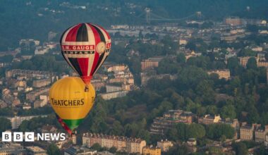 Two hot air balloons flying over Bristol. The city can be seen below, with the landscape featuring colourful properties and the Clifton Suspension Bridge as well as thick lines of trees.