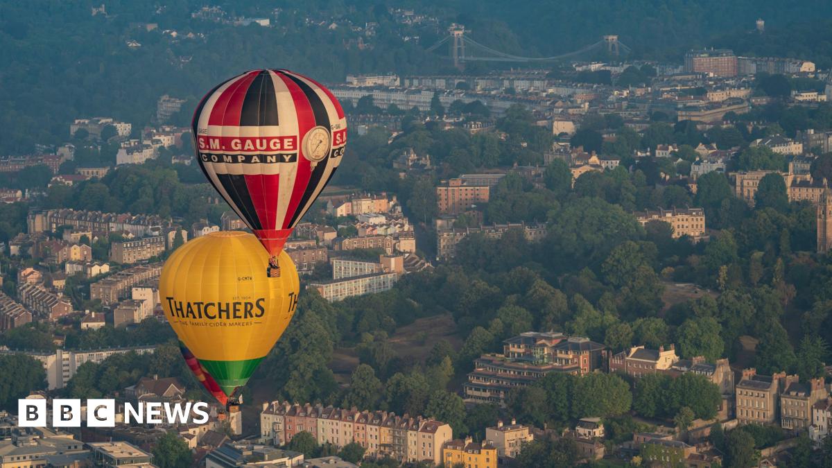 Two hot air balloons flying over Bristol. The city can be seen below, with the landscape featuring colourful properties and the Clifton Suspension Bridge as well as thick lines of trees.