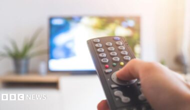 A stock image of someone watching TV. You can see a hand holding a black remote control, the background is out of focus but you can see the outline of a TV screen.