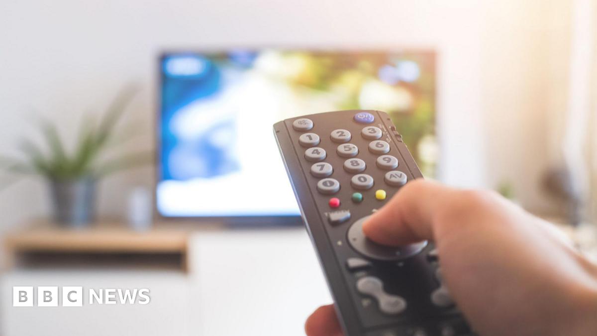 A stock image of someone watching TV. You can see a hand holding a black remote control, the background is out of focus but you can see the outline of a TV screen.