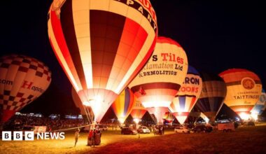 A number of hot air balloons illuminated at night time. They are stationary on a field, with crowds of people looking on.