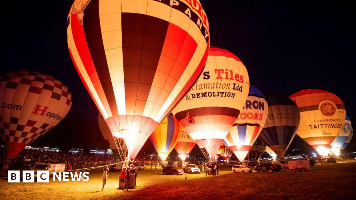 A number of hot air balloons illuminated at night time. They are stationary on a field, with crowds of people looking on.