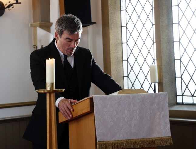 Cain struggles at a podium for Nate's funeral in Emmerdale