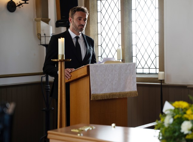 John stands at a podium at Nate's funeral in Emmerdale