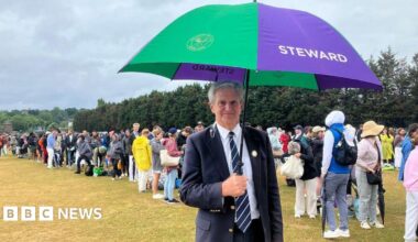 A man with grey hair wearing a navy blue suit and a striped white and navy blue tie is holding an umbrella. He is looking at the camera. Behind him, in a field, are queues of people, waiting to go into Wimbledon.