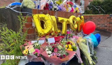 Bunches of flowers against a wall outside a house, with golden balloons spelling 'Dalton' above the flowers. Bunches of flowers on the top of the wall.