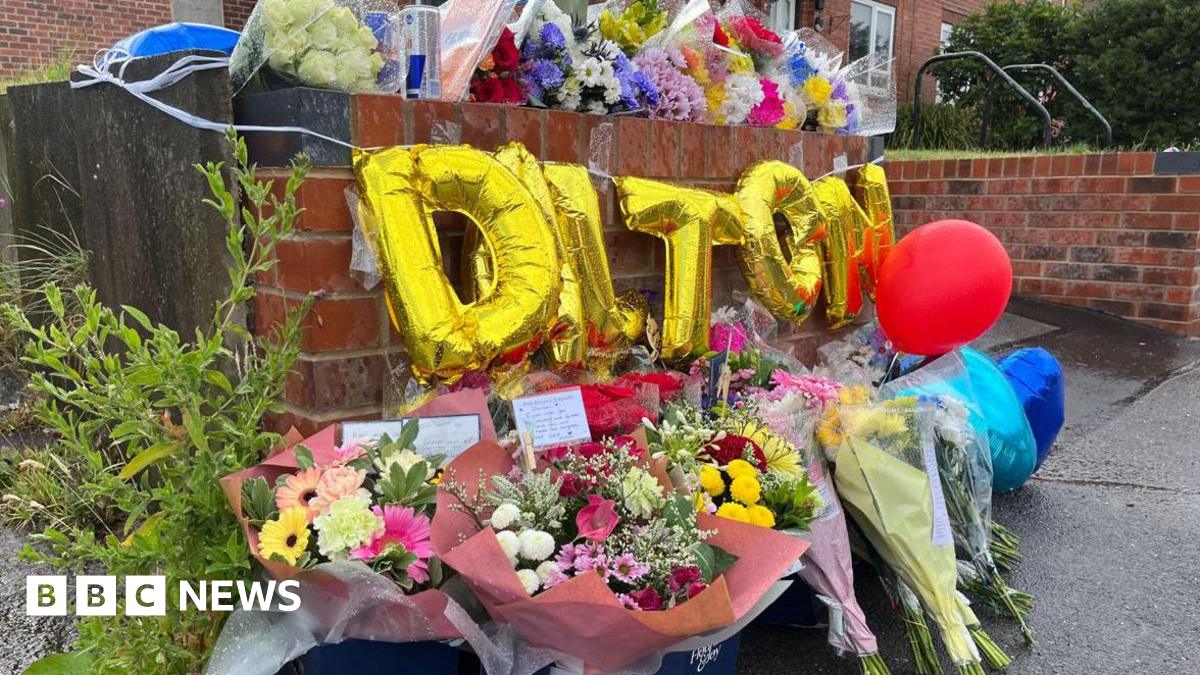 Bunches of flowers against a wall outside a house, with golden balloons spelling 'Dalton' above the flowers. Bunches of flowers on the top of the wall.