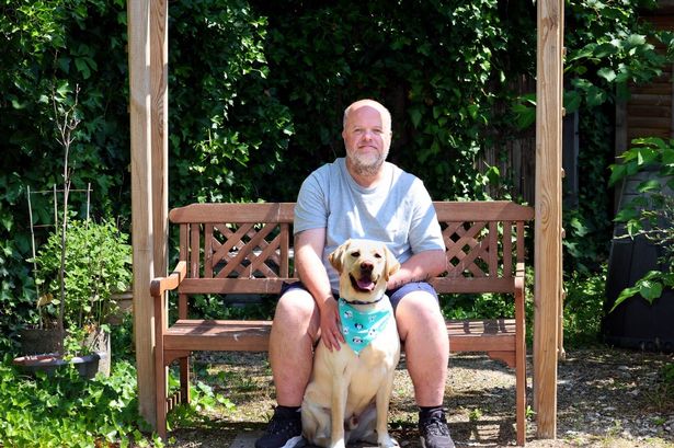 John Edgley with his guide dog Dodger sitting on a bench with ivy behind him. Dodger is sat in front of John wearing a bright blue neck scarf