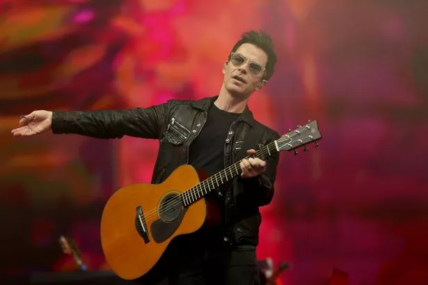 A man, dressed in black and with sunglasses, holds an acoustic guitar