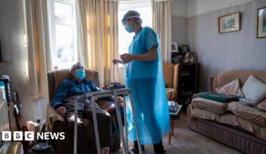 A healthcare worker standing and dressed in a blue gown chats to a housebound patient in his front room. The patient is sat down on a chair with a medical walking frame in front of him. He is wearing a mask. There is a sofa on the right of the picture and sun is coming through the windows behind.
