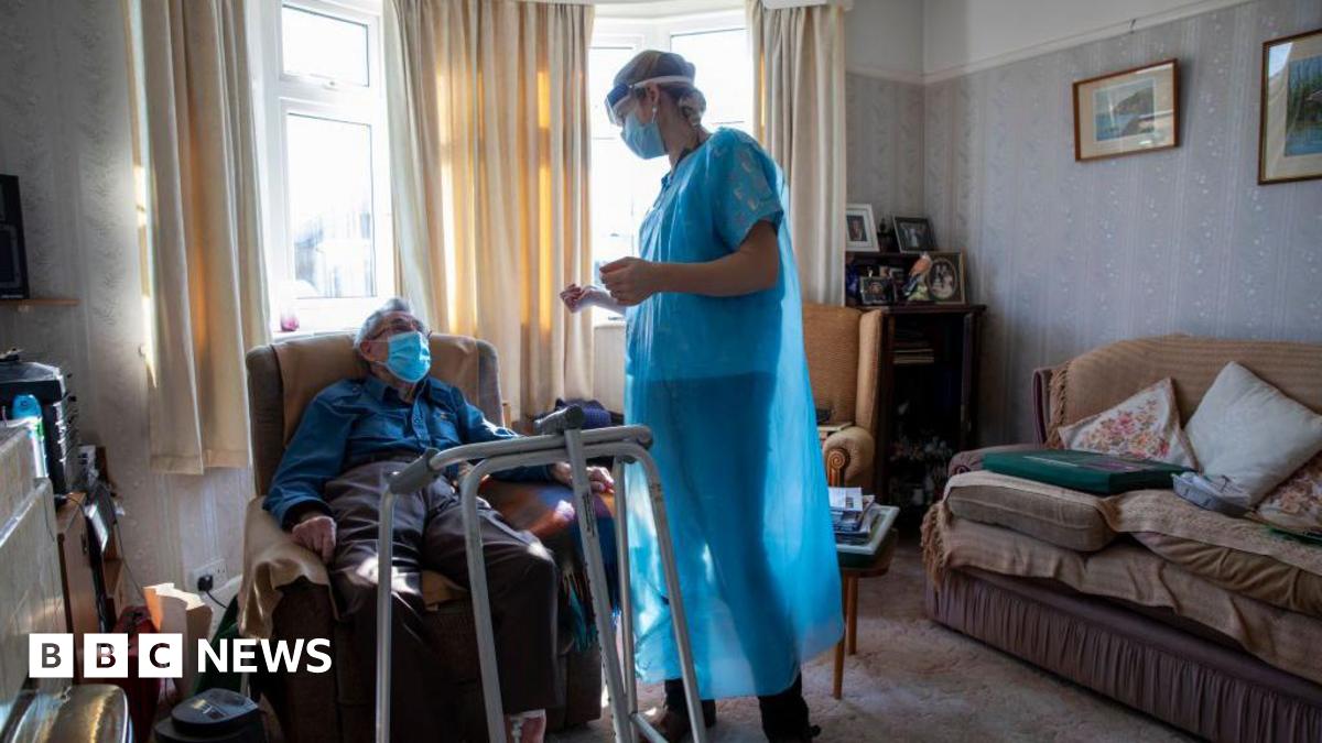 A healthcare worker standing and dressed in a blue gown chats to a housebound patient in his front room. The patient is sat down on a chair with a medical walking frame in front of him. He is wearing a mask. There is a sofa on the right of the picture and sun is coming through the windows behind.