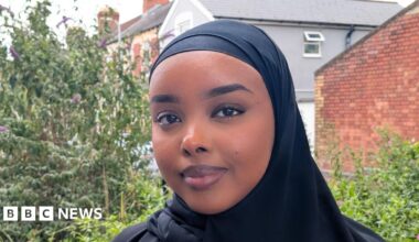 Munirah, a young Welsh-Somali woman stands on a street in Grangetown, Cardiff with trees and houses behind her. She's looking at the camera, smiling and wearing a black headscarf.