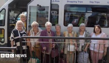 A group of older women dressed in casual clothes and smiling. They are standing next to a  small white bus.