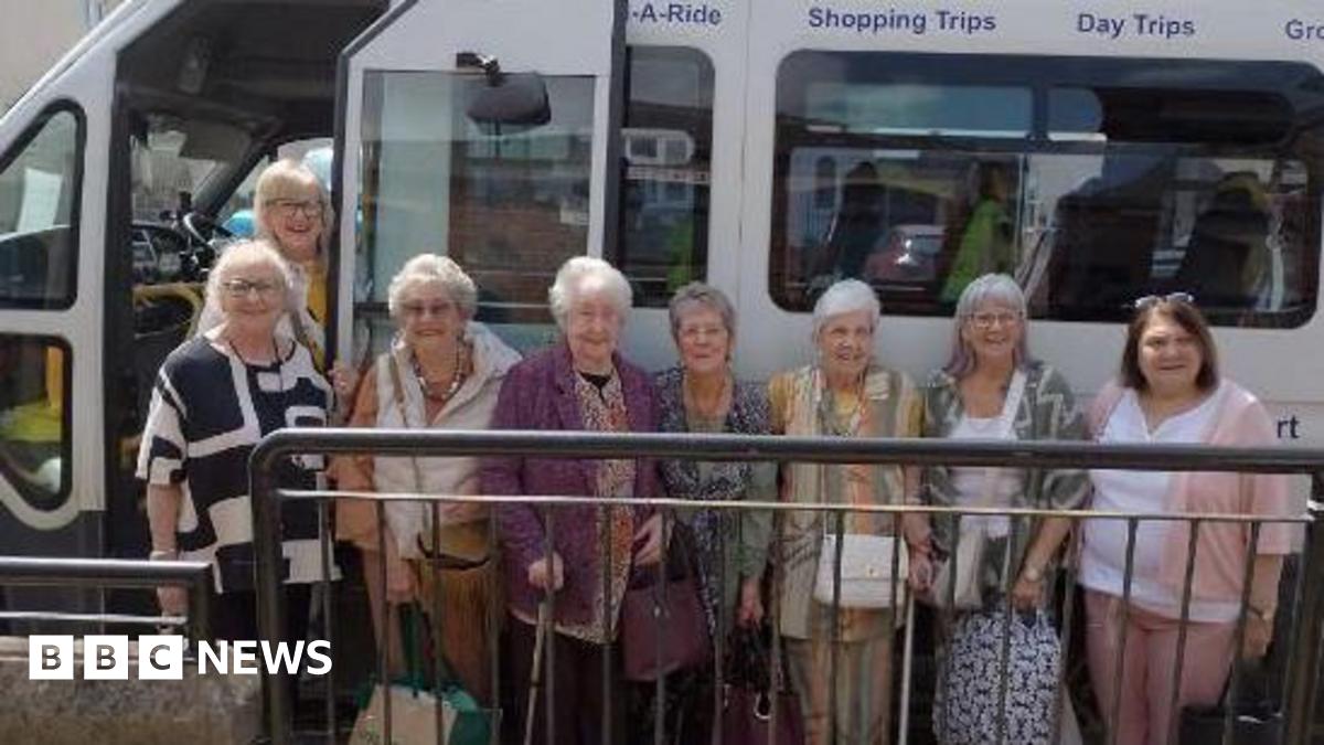 A group of older women dressed in casual clothes and smiling. They are standing next to a  small white bus.