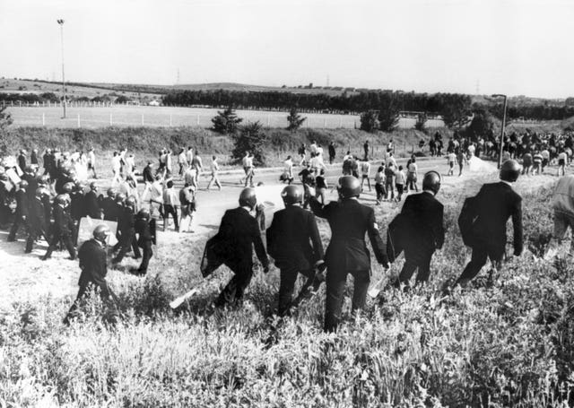 Black and white image of police in anti-riot gear escorting picketers away from their position near the Orgreave Coking Plant in South Yorkshire in 1984