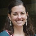 Kelly Monk, Ph.D., has brown hair pulled back in the front, a pink top and teal sweater, smiling in front of a building.