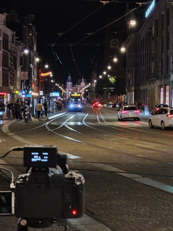 A nighttime city street with tram tracks, cars, and people. A camera on a tripod is in the foreground, and a tram approaches in the distance under streetlights. Shops and buildings line both sides of the street.