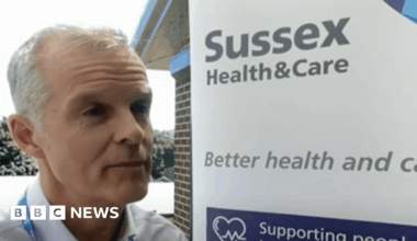 Stephen Lightfoot, a white man with silver hair, stands in front of a sign that reads "Sussex Health and Care - better health and care"