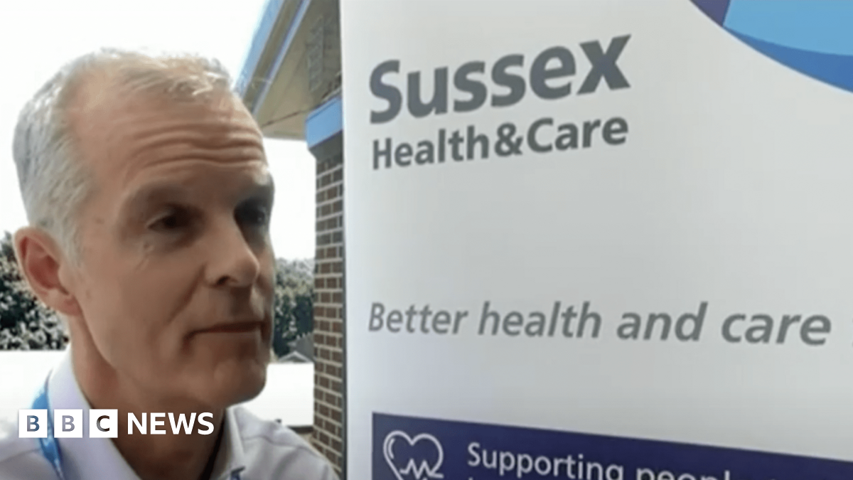 Stephen Lightfoot, a white man with silver hair, stands in front of a sign that reads "Sussex Health and Care - better health and care"