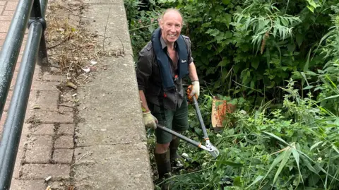 Sheffield City Council Overhead view of a middle-aged man stood in bushes on a river bank, snipping away with garden shears.