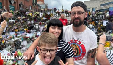 A man wearing a white Ozzy Osbourne t-shirt stands with his arm around a woman and a young boy. Behind them are hundreds of bouquets of flowers and tributes to Ozzy Osbourne at Black Sabbath Bridge.