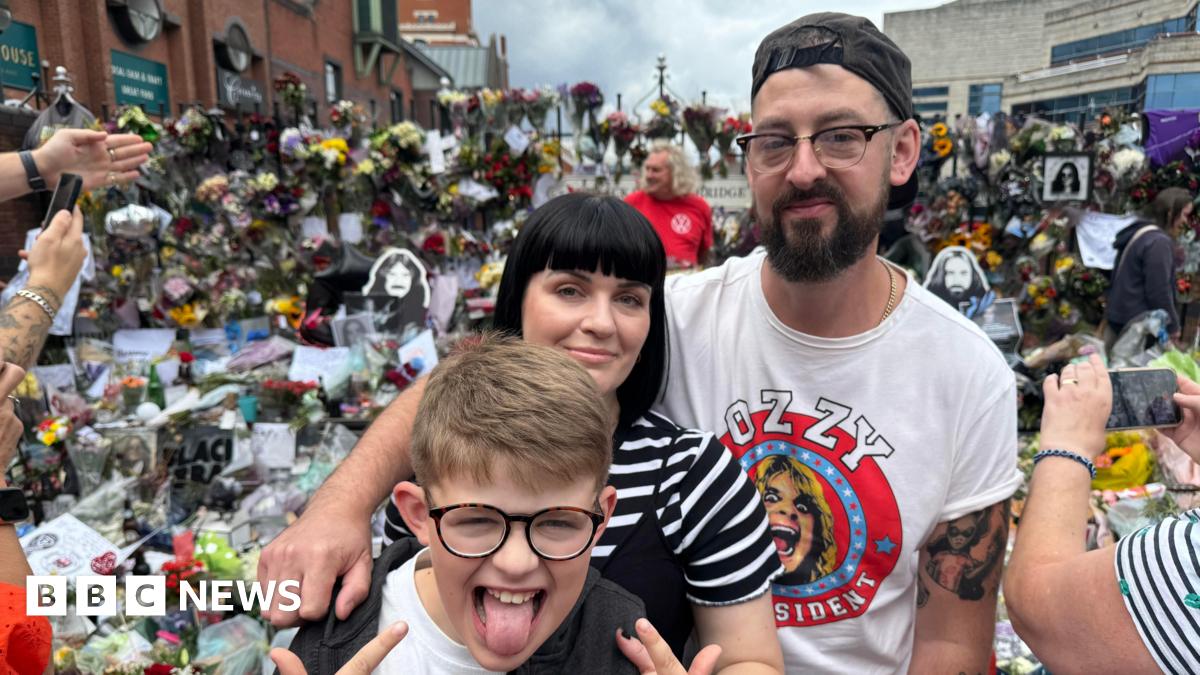 A man wearing a white Ozzy Osbourne t-shirt stands with his arm around a woman and a young boy. Behind them are hundreds of bouquets of flowers and tributes to Ozzy Osbourne at Black Sabbath Bridge.