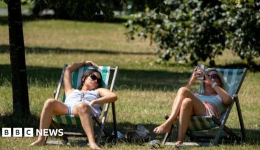 Two women sitting under tree shade on a noticeably hot day in a park in green and white striped deckchairs. The woman on the left has dark hair and sunglasses and appears to be asleep, with her right arm rest above her head on top of the deckchair. The woman on the right is blonde and is also wearing sunglasses and is looking at her phone.