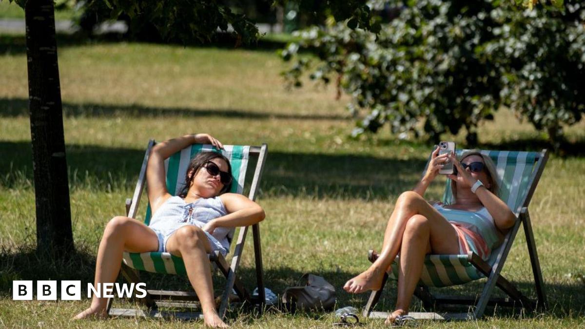 Two women sitting under tree shade on a noticeably hot day in a park in green and white striped deckchairs. The woman on the left has dark hair and sunglasses and appears to be asleep, with her right arm rest above her head on top of the deckchair. The woman on the right is blonde and is also wearing sunglasses and is looking at her phone.