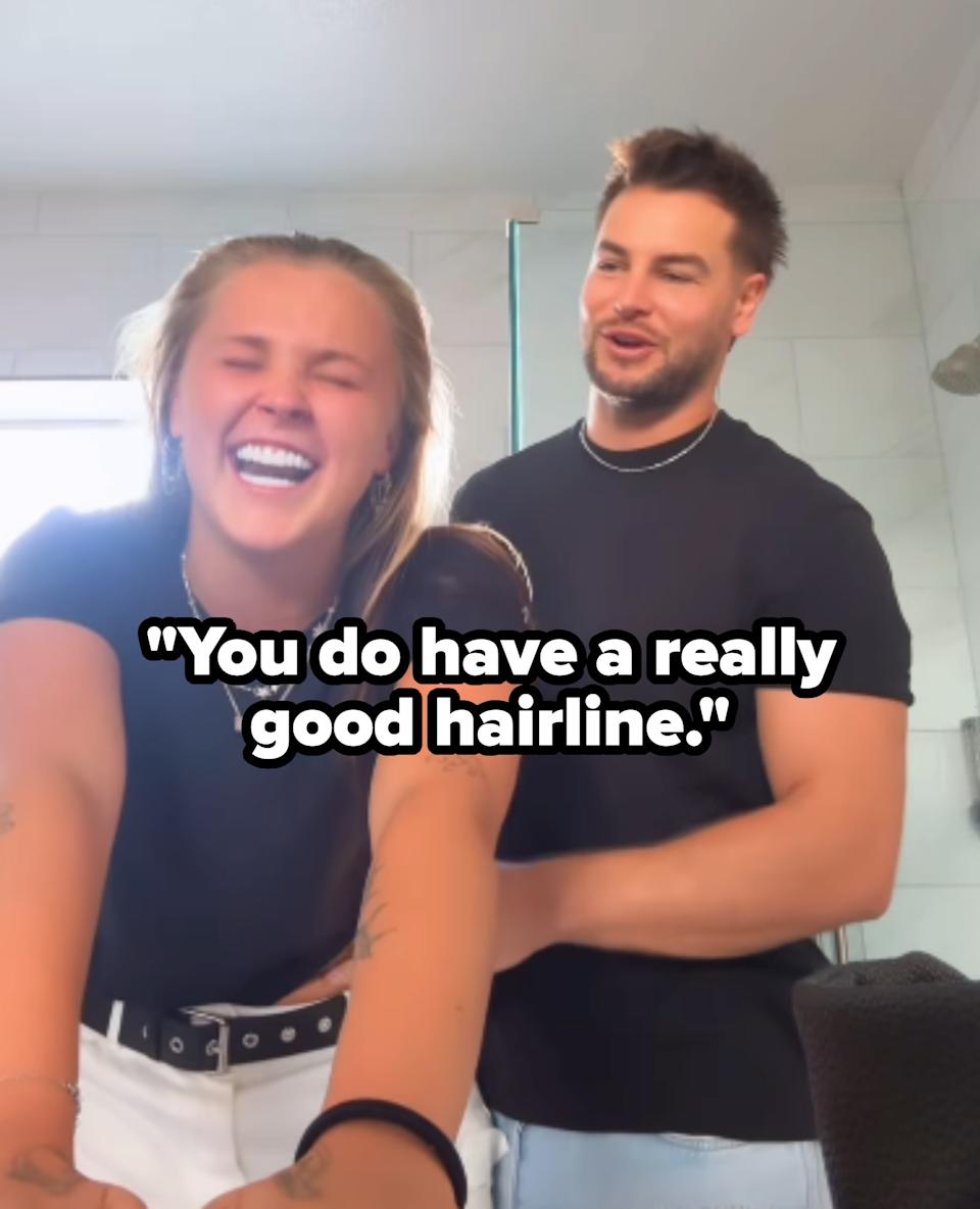 Two people smiling and laughing in a casual setting; one is leaning forward, and the other is standing behind them