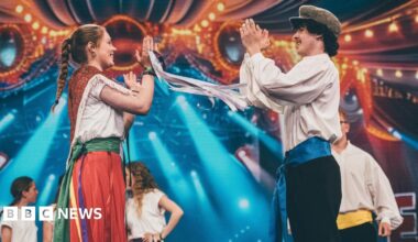 A group of young people folk dancing on the stage at the Eisteddfod in Pontypridd 2024. They are all in traditional outfits. There is a man and woman on the front of the stage with their hands in the air clapping whilst smiling at each other.