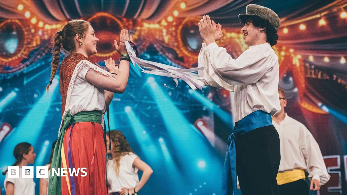 A group of young people folk dancing on the stage at the Eisteddfod in Pontypridd 2024. They are all in traditional outfits. There is a man and woman on the front of the stage with their hands in the air clapping whilst smiling at each other.