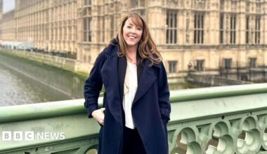 A woman with brown hair wearing a white shirt under a navy trench coat. She is standing in front of the Houses of Parliament.