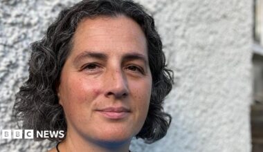 A woman with black and grey curly hair and brown eyes stands in front of a white wall