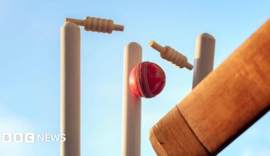 A cricket ball is hitting a wicket and the two bails are in the air in the process of being broken. A cricket bat can be seen in the bottom right corner. The blue sky behind the wicket.