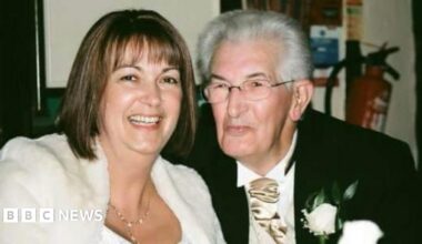 Lynda Costerd on the left with her father Geoff Toms on the right. They are attending a wedding. She has a fluffy white Faux fur shrug cardigan on, with a gold hanging necklace and brown bobbed hair with a fringe. He has a black suit jacket on with a gold tie and a cream flower in his pocket. Mr Toms is looking to the left of the camera and is smiling. He has glasses on.