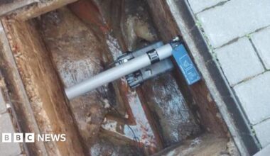 The view looking down a manhole showing a grey pipe connected to an electronics box, above an empty sewer pipe