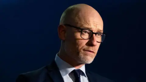 Getty Images Stephen Kinnock wearing dark glasses and a suit. There is a dark blue background behind him and he has a light shining on his face. He is looking off to the right. 