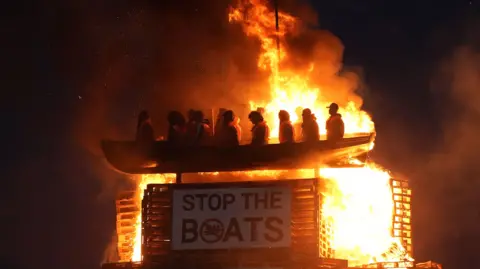 PA Media The picture shows a model of a small boat with figures inside in orange fluorescent vests. Below is a sign which says 'STOP THE BOATS'. It is on fire.